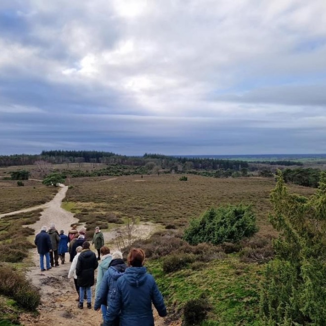 Groep mensen wandelt over een zandpad in een uitgestrekt natuurgebied met heuvels en bos op de achtergrond.