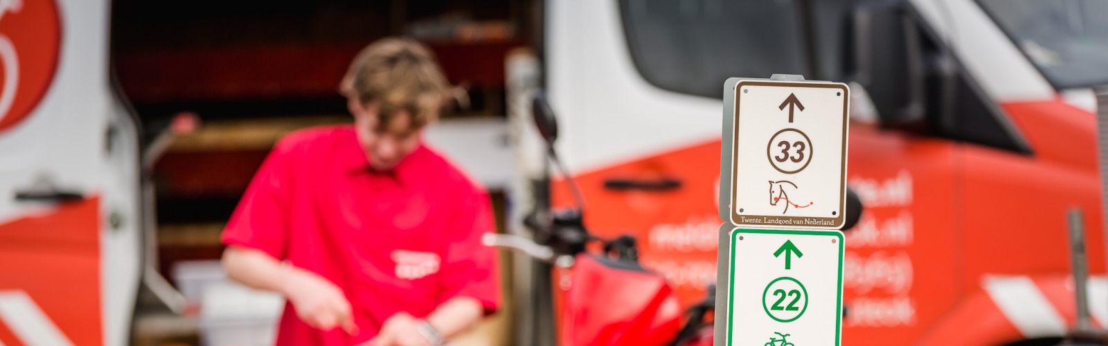 Bord met wandel-, fiets- en ruiterpadroutes bij een quad, met een persoon op de achtergrond in rood shirt.