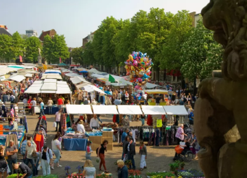 Drukke marktdag met kleurrijke kraampjes, mensen die winkelen, bomen en een stenen leeuwenbeeld op de voorgrond.