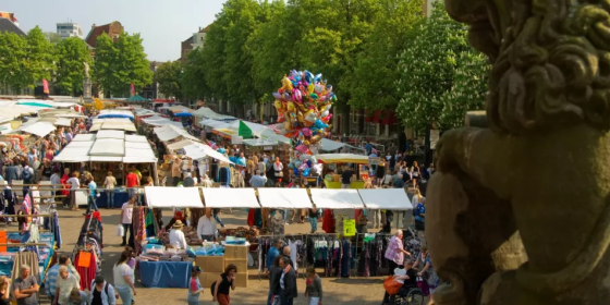 Drukke marktdag met kleurrijke kraampjes, mensen die winkelen, bomen en een stenen leeuwenbeeld op de voorgrond.