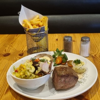 Een bord met biefstuk, frietjes in een mandje, gemengde salade, kruidenboter en groente op houten tafel.