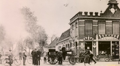 Historische foto van een straat met mensen, fietsen en een brandweerwagen voor een bakstenen gebouw.