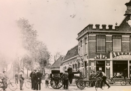 Historische foto van een straat met mensen, fietsen en een brandweerwagen voor een bakstenen gebouw.