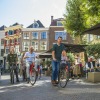 Mensen fietsen door een gezellige straat met terrasjes en historische gebouwen op een zonnige dag.