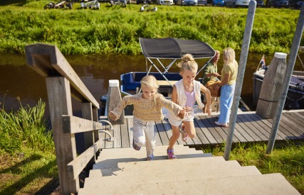 Twee meisjes rennen een trap op vanaf een steiger waar op de achtergrond een gezin bij een boot staat.