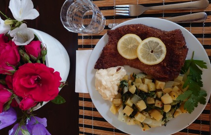 Een bord met gebakken schnitzel met citroen, aardappelsalade, peterselie en een bloemstuk ernaast.