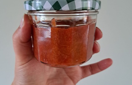 Hand houdt een glazen pot met roodbruine jam of saus en een geruit deksel tegen een lichte achtergrond.