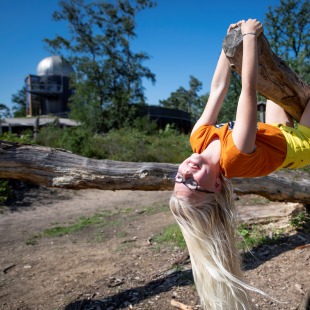 Een blond meisje met bril hangt lachend ondersteboven aan een boomtak in een zonnig natuurgebied.