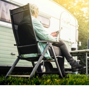 Een persoon zit ontspannen op een campingstoel voor een caravan met een tijdschrift, tafel en badmintonracket.