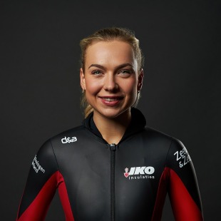 Portret van een glimlachende vrouwelijke sporter in een zwart-rood schaatspak met sponsorlogo’s.