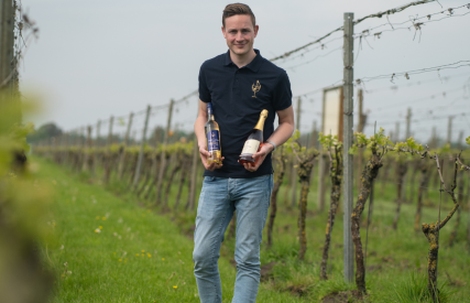 Man staat in een wijngaard met twee flessen wijn in zijn handen, gekleed in polo en spijkerbroek.