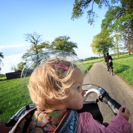 Een jong kind zit in een fietsstoeltje met een windscherm, fietsend over een landelijk pad in de zon.