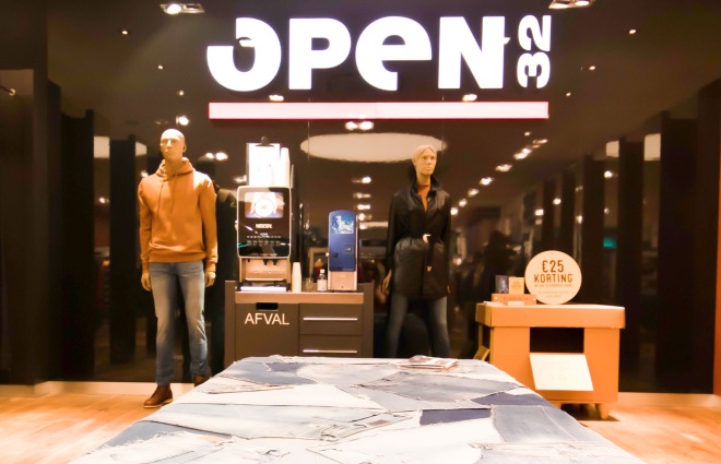 Een kledingwinkel met jeansdisplay, mannequins en een groot 'OPEN 32' bord op de achtergrond.