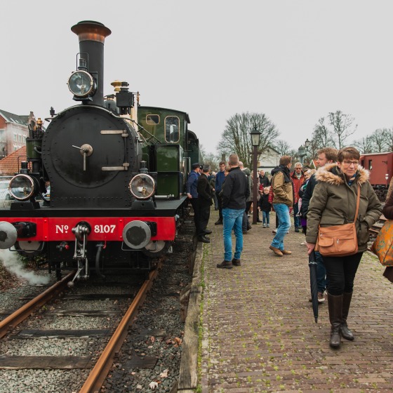 Menschenmenge steht neben einer historischen Dampflok an einem Bahngleis an einem bewölkten Tag.