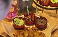 Een houten tafel met gefrituurde bakbananen, guacamole, bonenpuree en salsa in kleine kommetjes.