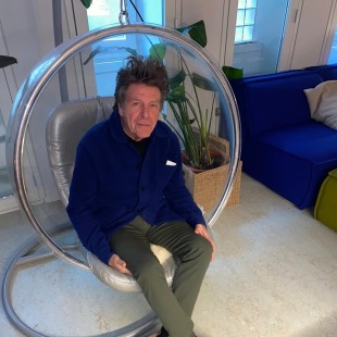 Man zit ontspannen in een ronde, hangende stoel in een moderne kamer met planten en een blauwe bank.