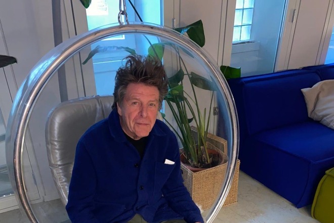 Man sits relaxed in a circular hanging chair in a modern room with plants and a blue couch visible.
