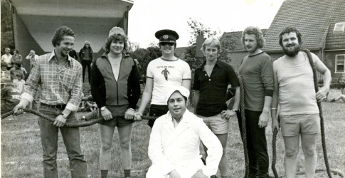 Zes mannen poseren met een touw buiten op het gras, een man in witte jas hurkt voor hen, jaren 70.