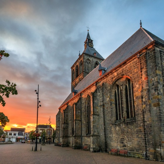 Historische kerk op een leeg plein bij zonsondergang, met dramatische lucht en kleurrijke bloemen links.