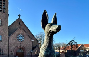 Beeld van een zittende hond met grote oren op een sokkel, op een plein bij een kerk en huizen, zonnige dag.