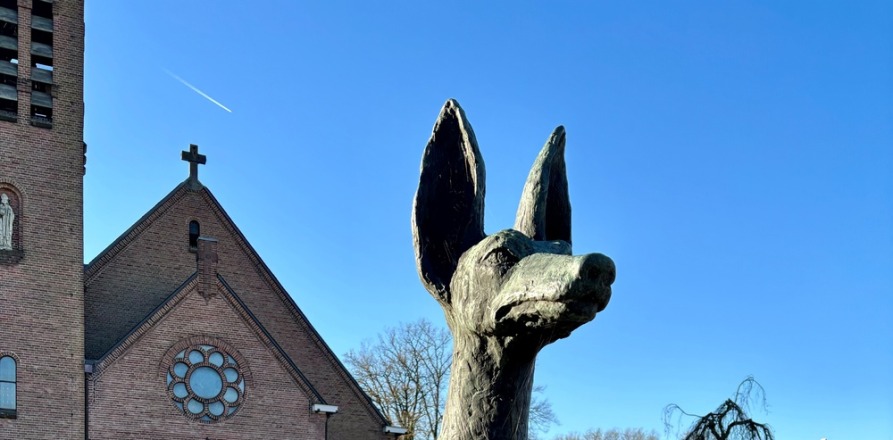Beeld van een zittende hond met grote oren op een sokkel, op een plein bij een kerk en huizen, zonnige dag.