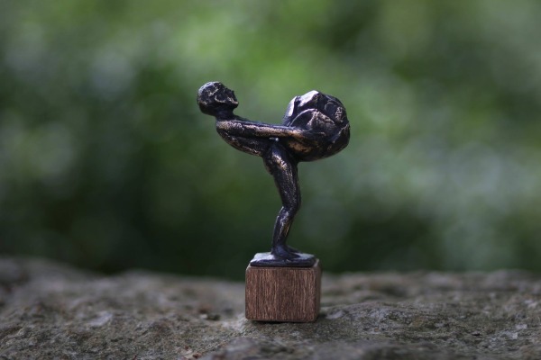 Bronzen miniatuurbeeldje van een persoon die een grote kool vasthoudt, geplaatst op een houten sokkel.