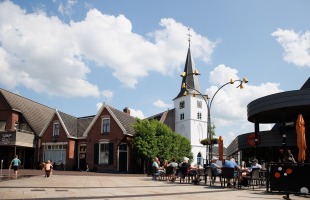 Gezellige terrassen op een dorpsplein met een witte kerktoren en blauwe lucht met wolken op de achtergrond.