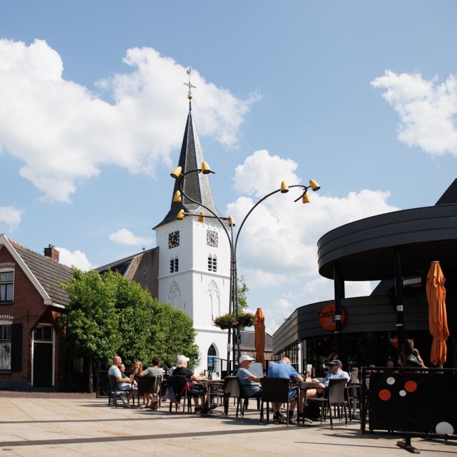 Gemütliche Sitzplätze im Freien auf einem Dorfplatz mit weißem Kirchturm und blauem Himmel mit Wolken.