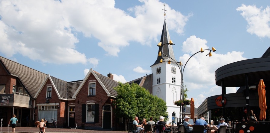 Gezellige terrassen op een dorpsplein met een witte kerktoren en blauwe lucht met wolken op de achtergrond.