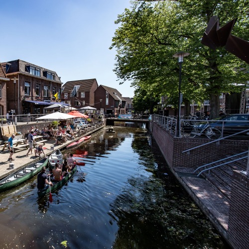 Mensen genieten van kanoën en terrassen langs een kanaal in een zonnige, schilderachtige stad in Nederland.