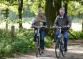 Twee volwassenen fietsen samen op een bosrijk pad, omgeven door groene bomen en zonnig weer.