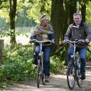 Twee volwassenen fietsen samen op een bosrijk pad, omgeven door groene bomen en zonnig weer.