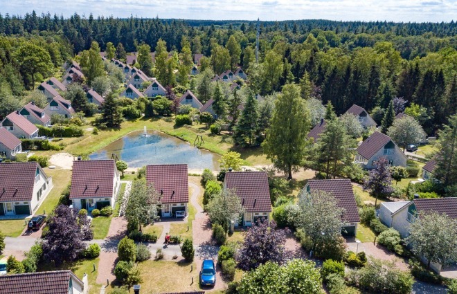 Luchtfoto van een vakantiepark in een bosrijk gebied met huisjes, vijver en veel groene bomen.