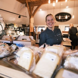 Vriendelijke medewerkster lacht achter een toonbank vol verpakte kazen in een gezellige kaaswinkel.