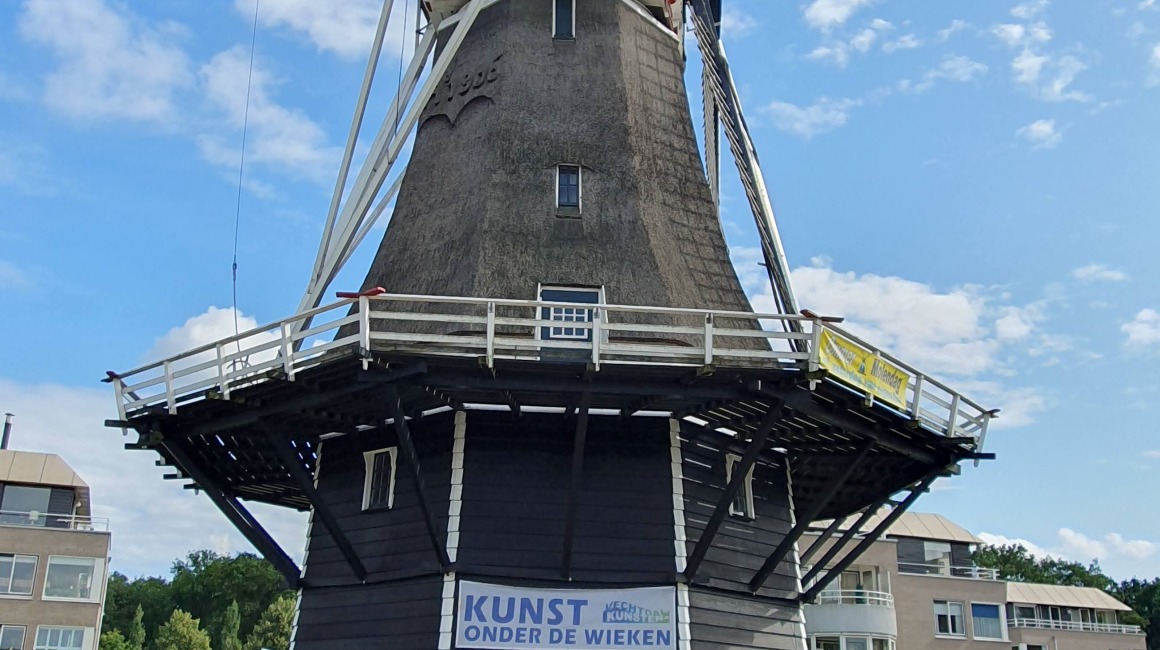 Historic Dutch windmill with art exhibition entrance, blue sky, and surrounding modern residential buildings.
