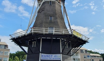 Historische Nederlandse windmolen met kunsttentoonstelling, blauwe lucht en omliggende moderne gebouwen.