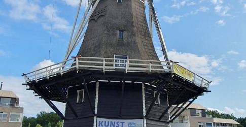 Historische Nederlandse windmolen met kunsttentoonstelling, blauwe lucht en omliggende moderne gebouwen.