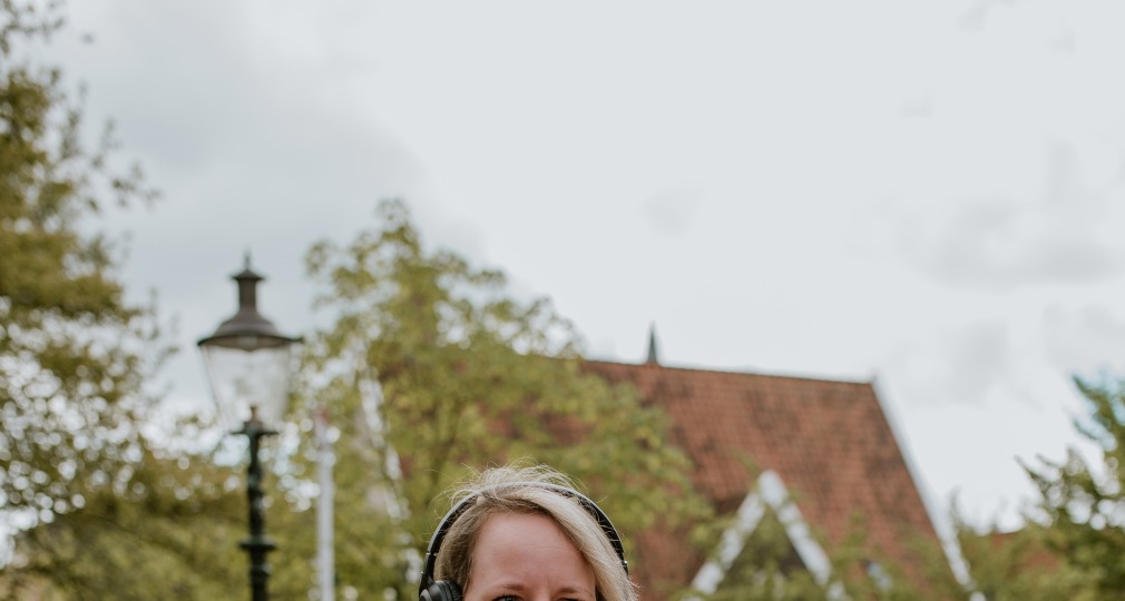 Vrouw met een zwart leren jack en koptelefoon wandelt buiten op straat met een telefoon in haar hand.