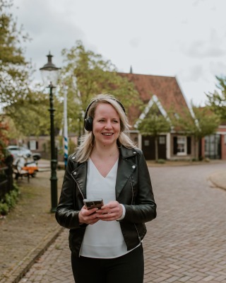 Vrouw met een zwart leren jack en koptelefoon wandelt buiten op straat met een telefoon in haar hand.