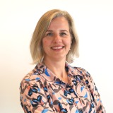 Portret van een glimlachende vrouw met blond haar in een kleurrijke blouse tegen een witte achtergrond.