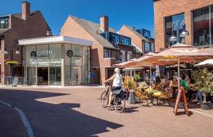 Ein Radfahrer fährt an einem modernen Einkaufsviertel und einem Straßencafé mit Sonnenschirmen vorbei.