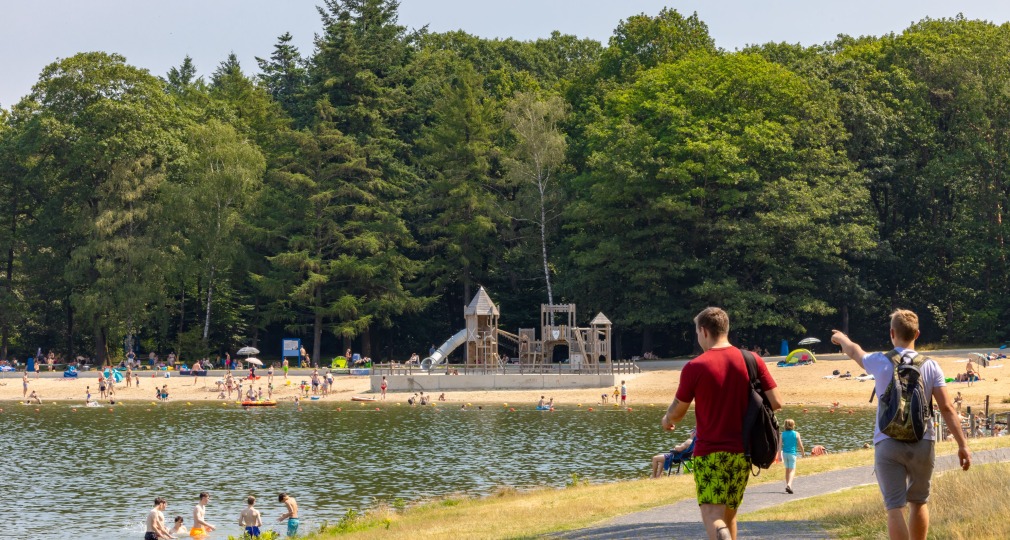 Twee mannen lopen langs een meer met strand en een speeltuin, omringd door bomen op een zonnige dag.