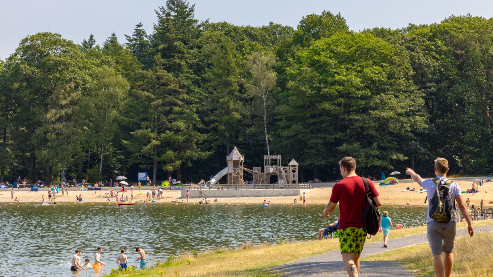 Twee mannen lopen langs een meer met strand en een speeltuin, omringd door bomen op een zonnige dag.