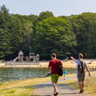 Twee mannen lopen langs een meer met strand en een speeltuin, omringd door bomen op een zonnige dag.