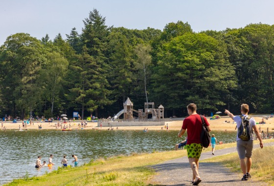 Twee mannen lopen langs een meer met strand en een speeltuin, omringd door bomen op een zonnige dag.