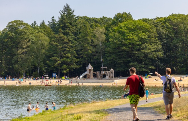 Twee mannen lopen langs een meer met strand en een speeltuin, omringd door bomen op een zonnige dag.