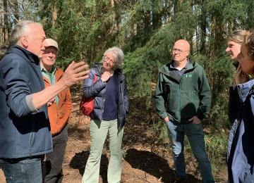 Groep volwassenen luistert aandachtig naar een gids tijdens een boswandeling op een zonnige dag.
