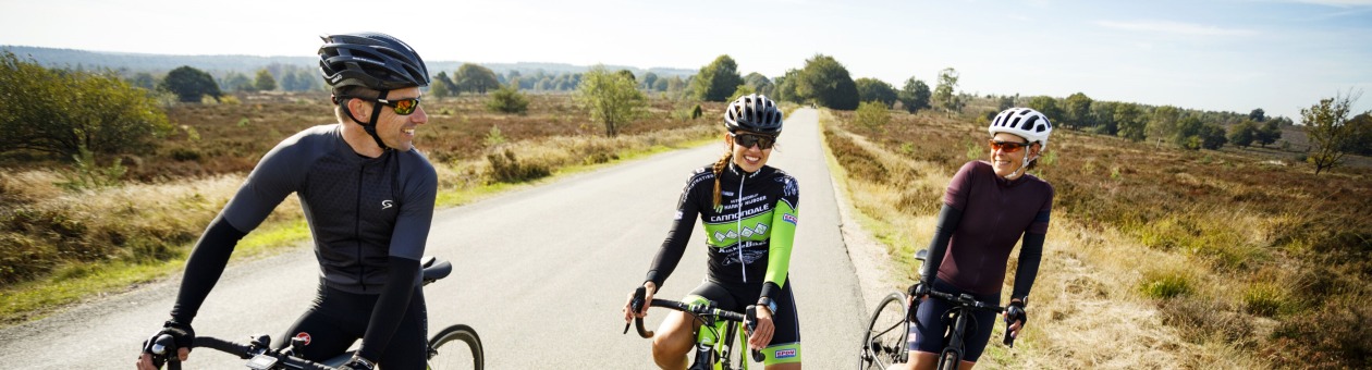 Drie wielrenners in sportkleding maken een praatje op een landelijke weg op een zonnige dag.