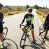 Drie wielrenners in sportkleding maken een praatje op een landelijke weg op een zonnige dag.