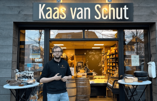 Man staat voor winkel 'Kaas van Schut' met kazen op tafels buiten en uitgestalde kaasproducten binnen.